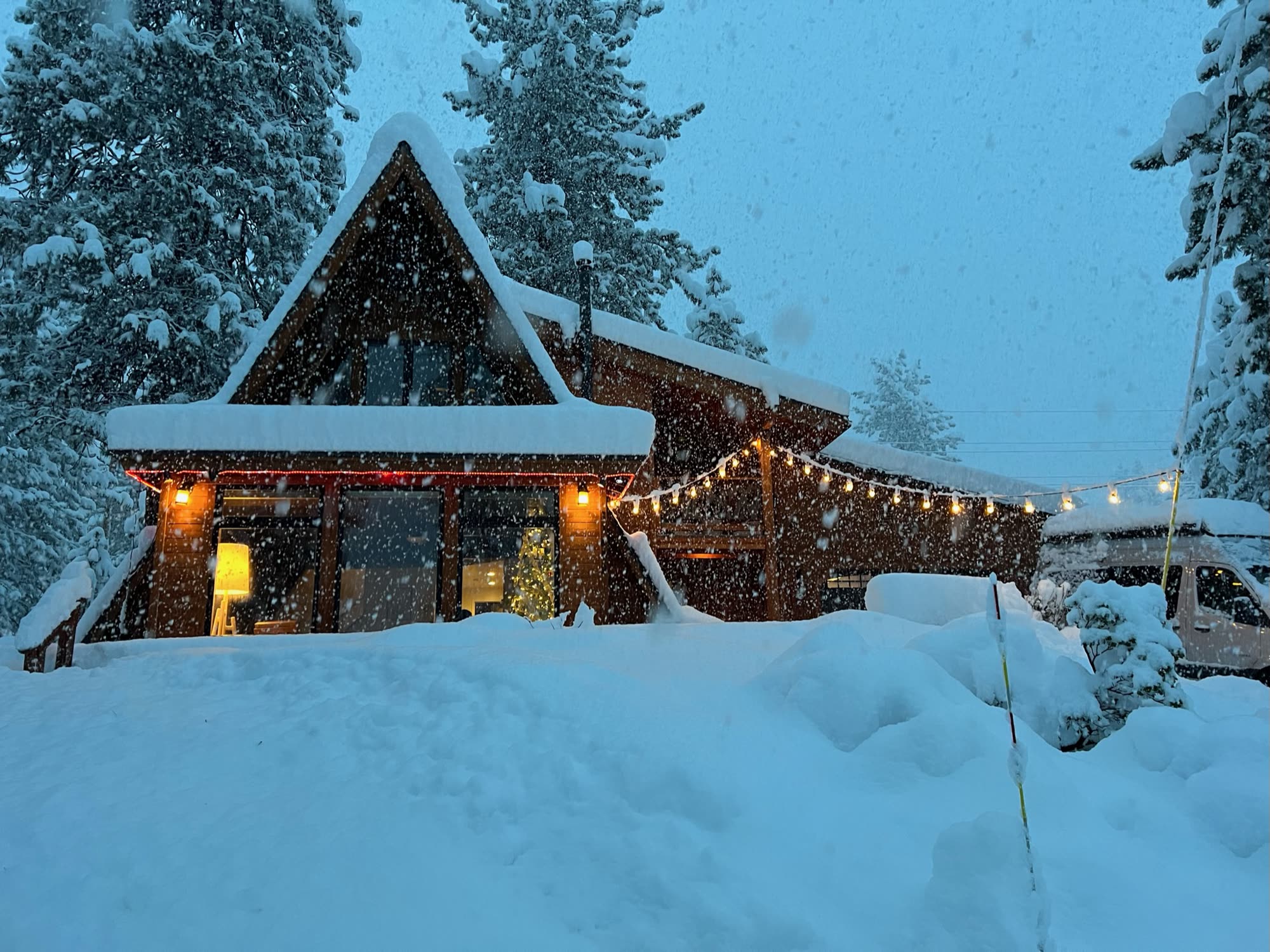 The A-frame in falling snow at dusk, windows glowing and string lights on