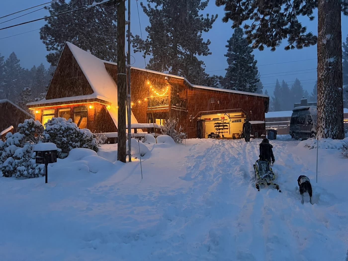 Walking home to the lit A-frame through fresh snow at dusk, the dog alongside